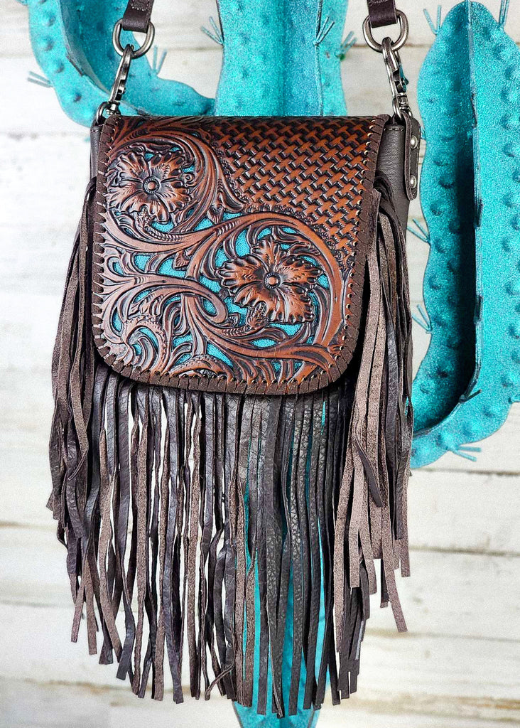 Western cowgirl purse with coffee floral and basket weaved tooling and matching leather whip stitch. Underneath floral tooling is a turquoise base to make floral design pop. Removable shoulder strap clasp are visible. Purse is hanging from a blue cactus metal rack.