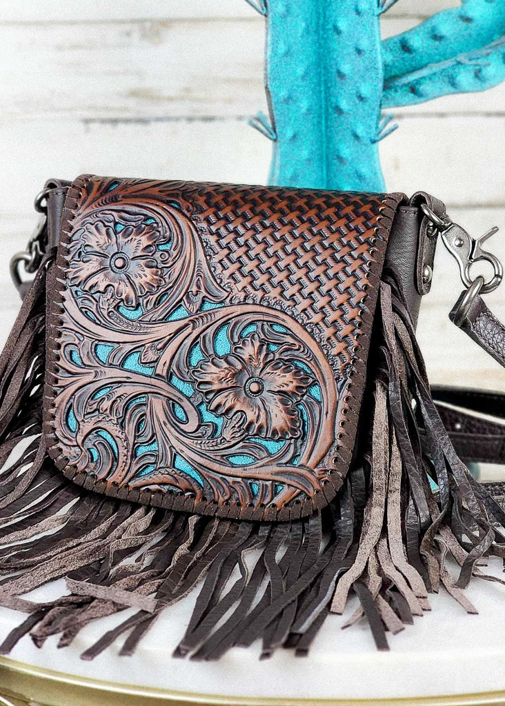 Western cowgirl purse with brown floral and basket weaved tooling and matching leather whip stitch. Underneath floral tooling is a cream base to make floral design pop. Removable shoulder strap clasp are visible. Purse sits on marble table top.  