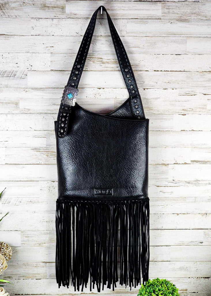 Brown wrangle hobo with fringe handing up against a wooden wall. Silver studded adjustable shoulder strap with turquoise concho. 