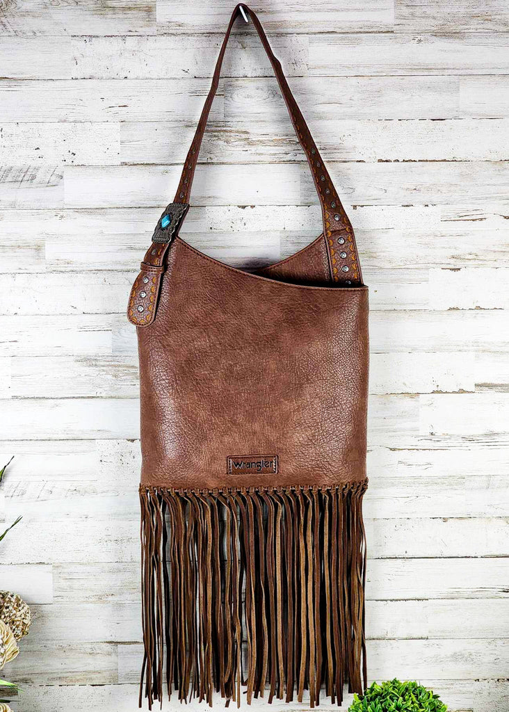 Brown wrangle hobo with fringe handing up against a wooden wall. Silver studded adjustable shoulder strap with turquoise concho. 