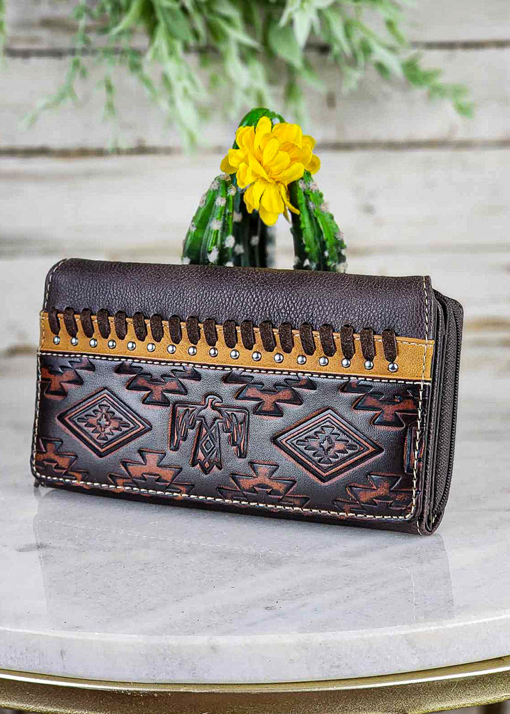 Southwestern wallet with coffee colored leather and contrasting lighter color where embossments are. Thunderbirds and tribal design embossments. Lighter color leather with silver studs and coffee whip stitch. Wallet leans against a cactus on a marble stool.