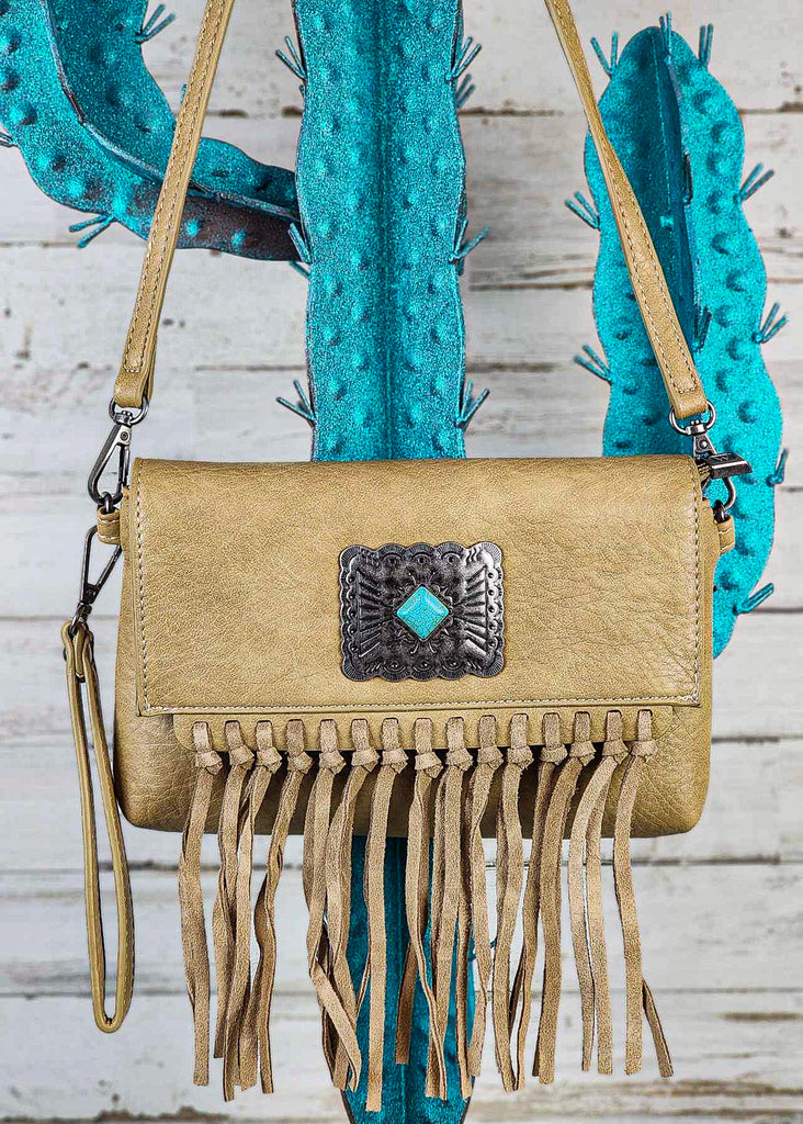 Tan western clutch with turquoise stone concho. Flap has hand tied fringe. Crossbody strap attached and hangs from blue cactus rack. Wristlet falls from side of clutch. 