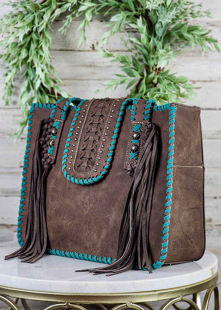 Western concealed carry tote bag with turquoise whip stitch, coffee fringe and silver concho studs. Snap cross over to close the bag has silver studs, braiding and turquoise whip stitch. Tote sits on marble stool.