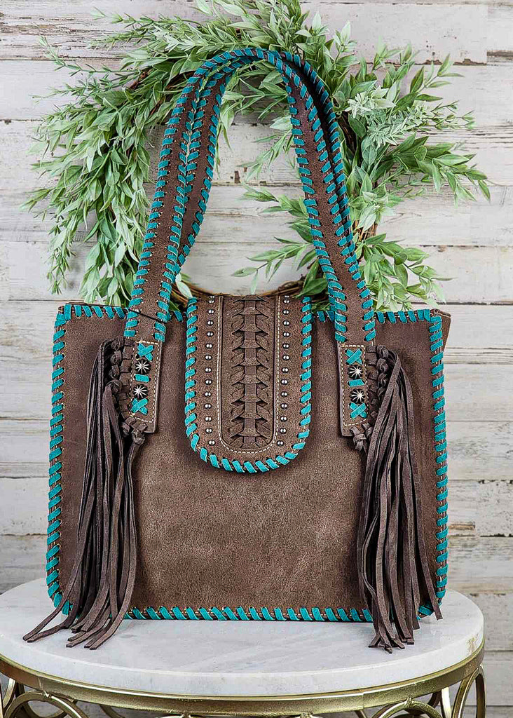 Western concealed carry tote bag with turquoise whip stitch, coffee fringe and silver concho studs. Snap cross over to close the bag has silver studs, braiding and turquoise whip stitch. Tote sits on marble stool. 