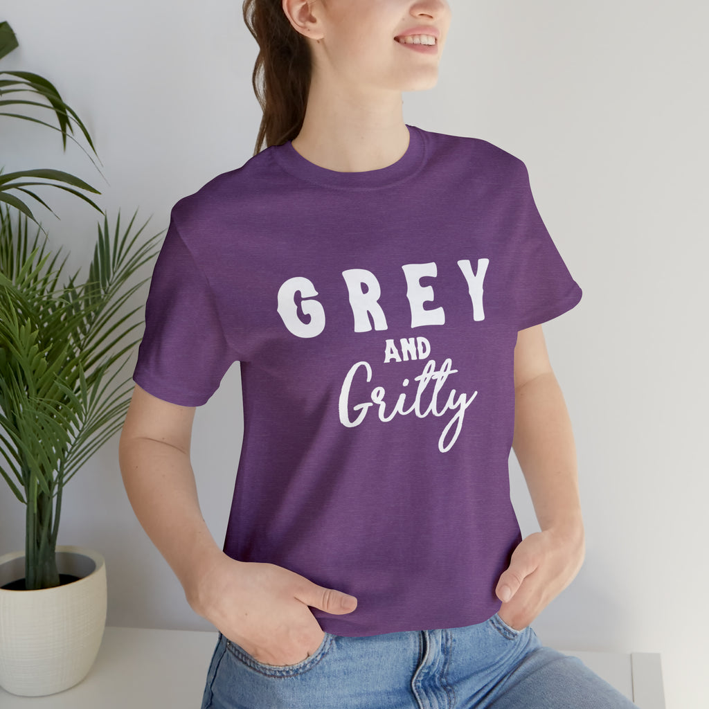 Purple short sleeve with white lettering reading "Grey and Gritty". Model wears this with jeans and her hands tucked into the pockets.