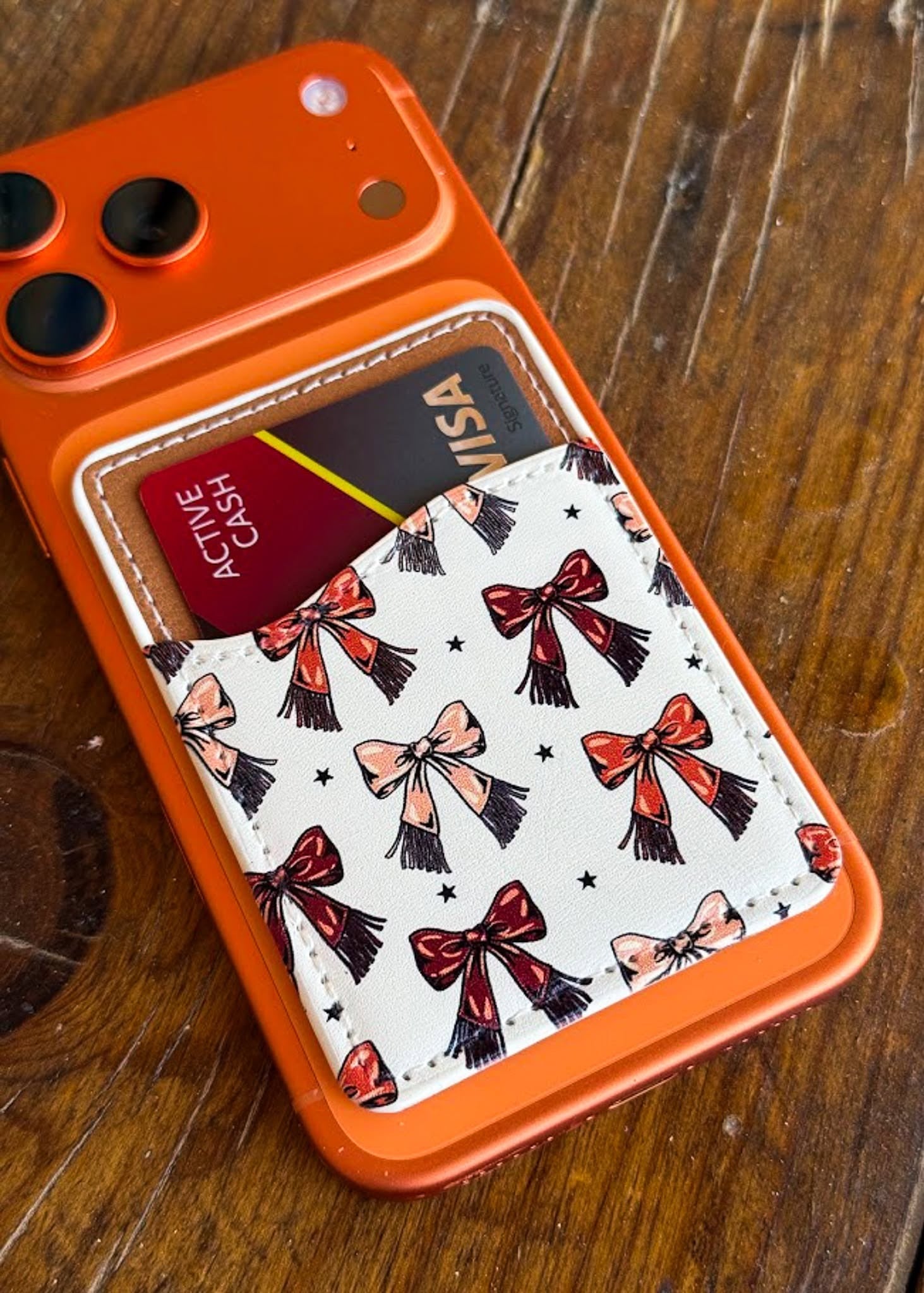 An orange phone case with a card holder pocket featuring a white background and a bow pattern, shown on a wooden surface.