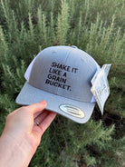 A light gray snapback cap with black embroidered text reading 'SHAKE IT LIKE A GRAIN BUCKET.' held by a hand against a background of green shrubs.