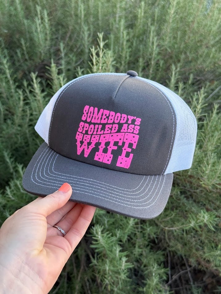 A hand holding a dark gray and white trucker hat with pink text that reads 'SOMEBODY'S SPOILED ASS GIFTED INTELLIGENT WOMAN' against a background of green shrubs.