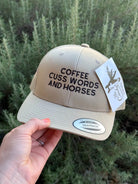 A beige snapback cap with black embroidered text reading 'COFFEE CUSS WORDS AND HORSES' is held by a hand against a background of green shrubs. A white hangtag and a silver visor sticker are attached.