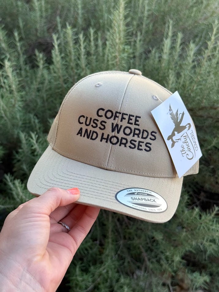 A beige snapback cap with black embroidered text reading 'COFFEE CUSS WORDS AND HORSES' is held by a hand against a background of green shrubs. A white hangtag and a silver visor sticker are attached.
