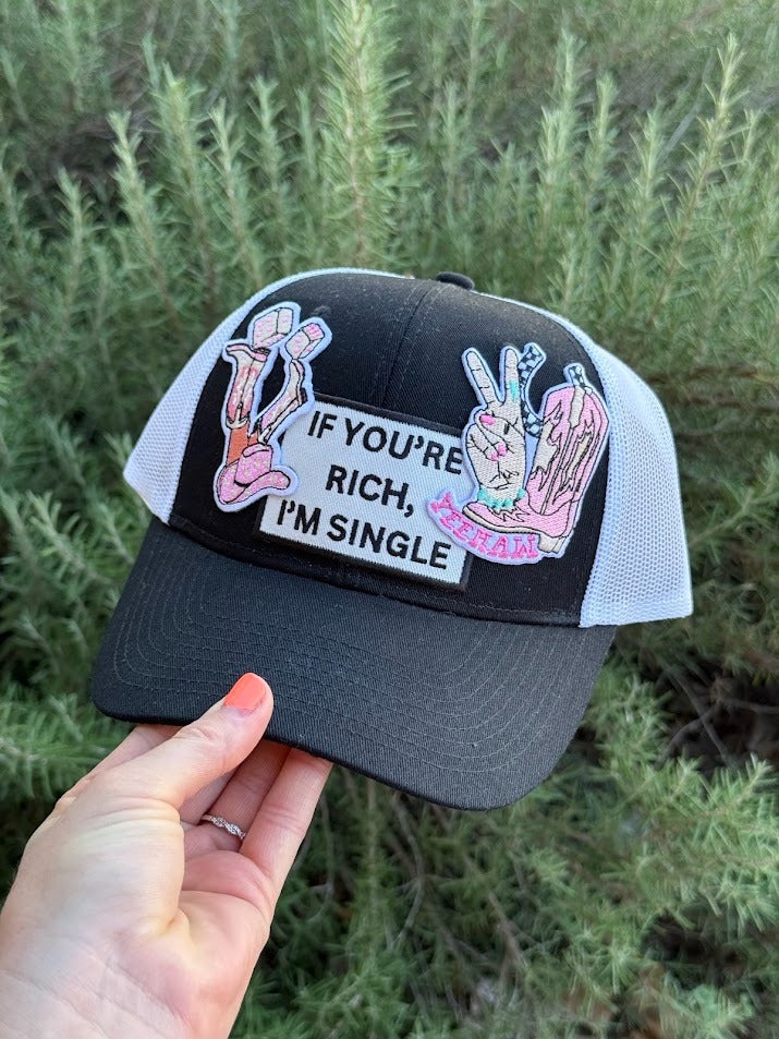 A hand holds a black and white trucker hat with a graphic patch that reads 'IF YOU'RE RICH, I'M SINGLE' against a background of green plants.