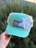 A mint green and beige trucker hat with a patch reading 'WORKING CATTLE BUILDS VOCABULARY' held by a hand against a background of green plants. A white hangtag is attached to the side.