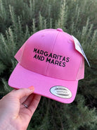 A pink trucker-style cap with black embroidered text reading 'Margaritas and Mares' is held by a hand against a blurred green outdoor background. The cap has a structured front panel, black mesh side panels, a white hangtag, and a Snapback sticker on the brim.