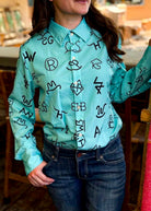 A woman wearing a mint green button-down shirt with a black graphic monogram pattern, paired with dark blue jeans, in a warm indoor setting.