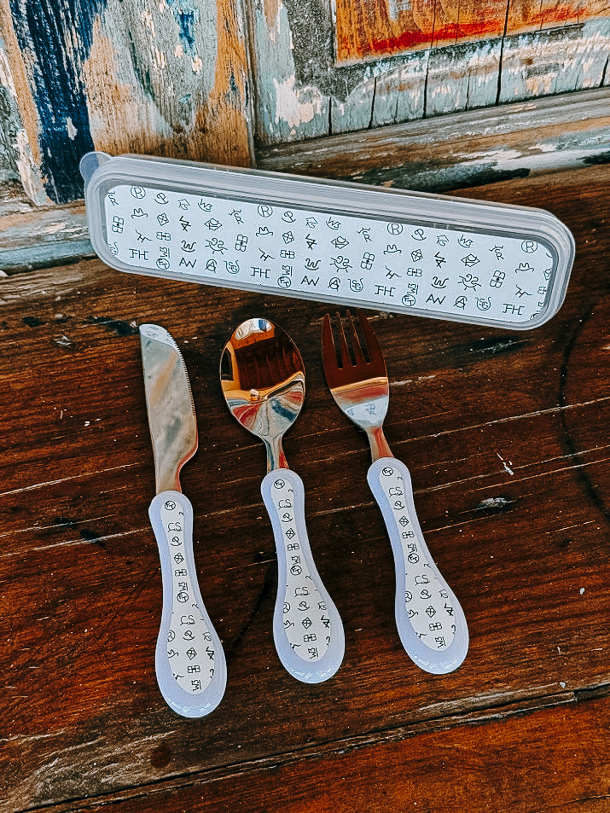 Three personalized stainless steel cutlery pieces with white handles printed with black monogram letters, displayed on a rustic wooden table next to their matching storage case.