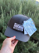 A dark gray trucker hat held by a hand, featuring a white embroidered patch that reads 'I JUST WANT TO DRINK MARGARITAS & RIDE MY HORSE' and a light blue hangtag with the same text, set against a background of green plants.