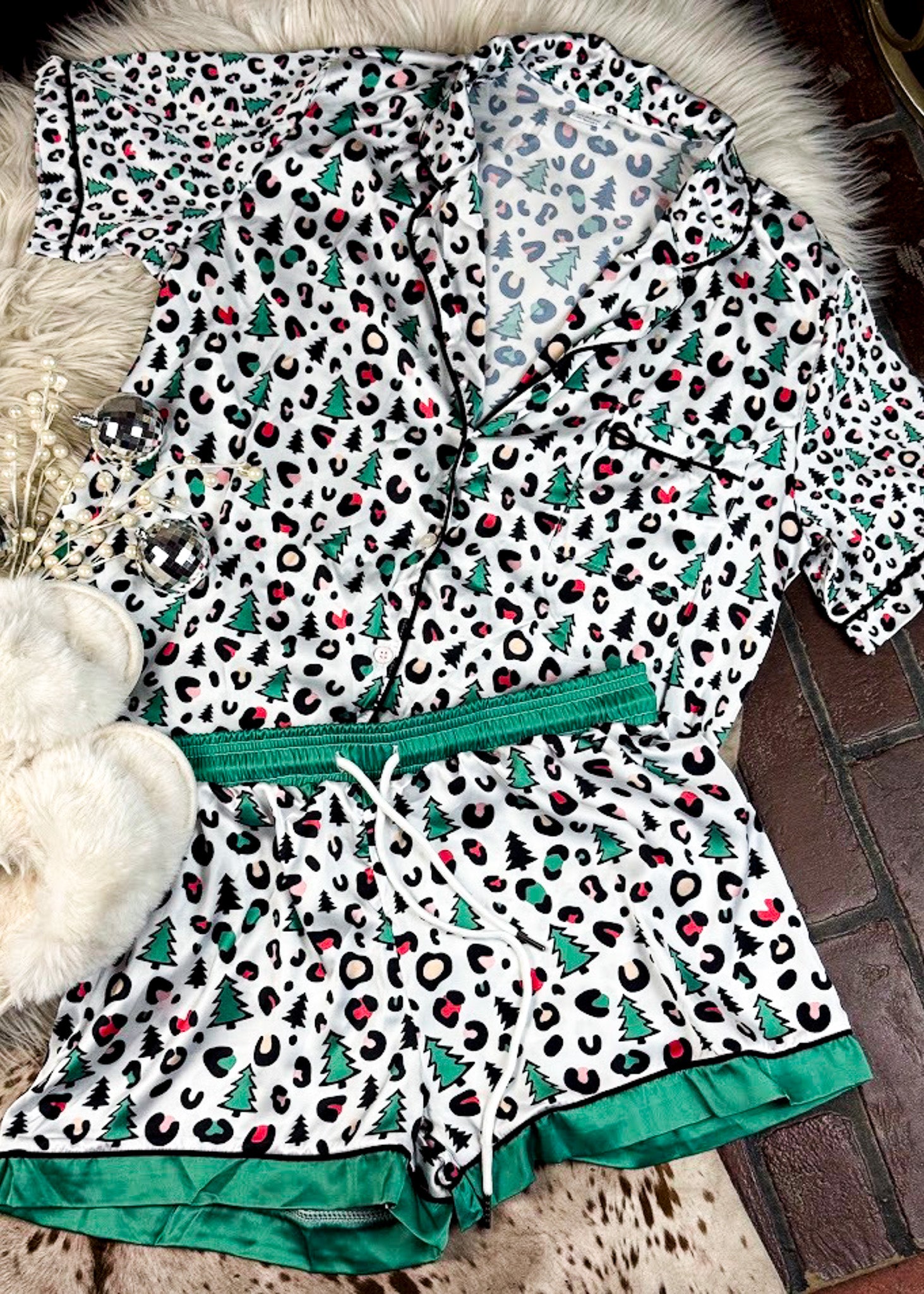leopard print and green with christmas trees silky pajamas, two piece set with shorts, laying on a white fluffy rug with white fluffy slippers 