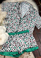 leopard print and green with christmas trees silky pajamas, two piece set with shorts, laying on a white fluffy rug with white fluffy slippers 