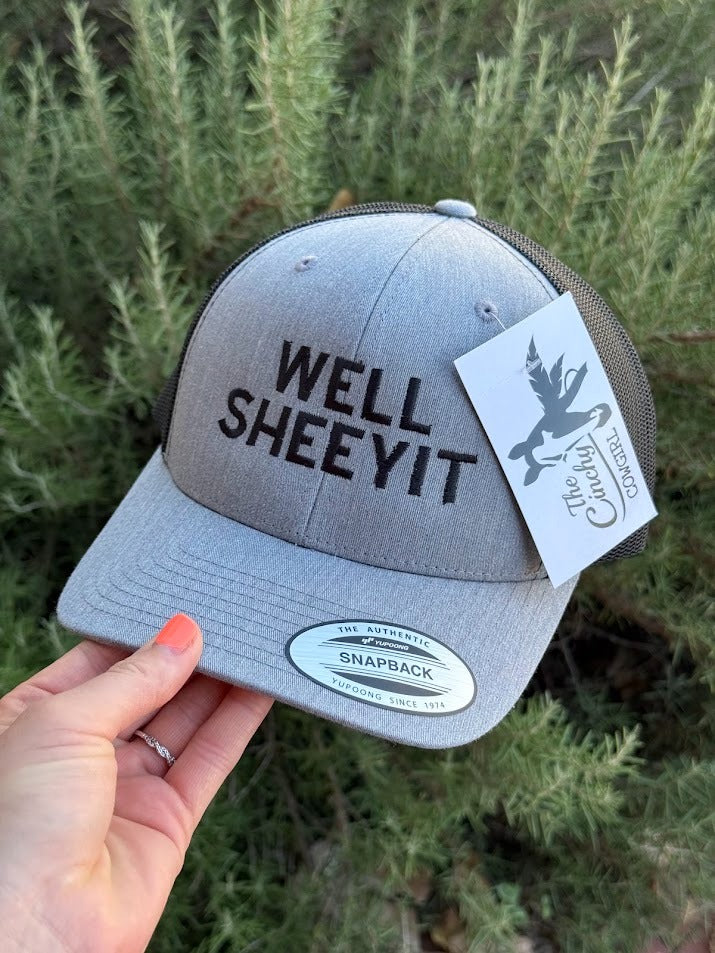 A light gray snapback cap with 'WELL SHEYIT' printed in black on the front, held by a hand against a background of green pine branches. A white paper tag from 'The Gathering Place' is attached to the side.