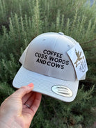 A light gray snapback cap with black embroidered text reading 'COFFEE CUSS WORDS AND COWS' is held by a hand in front of green foliage. The cap has a white hangtag with a horse logo and a visible Snapback brand sticker on the brim.