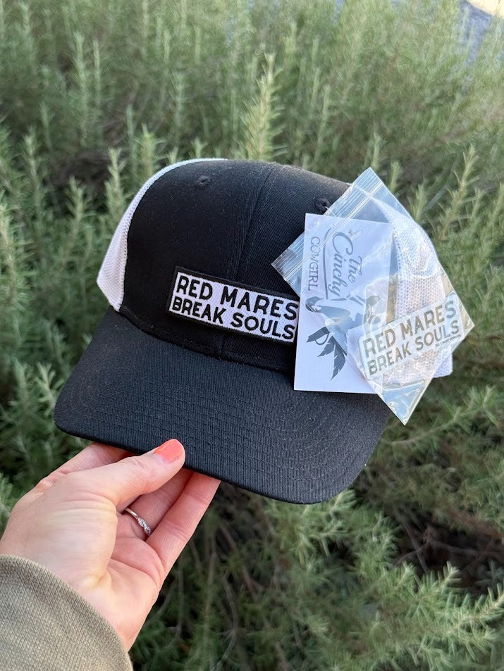 A black and white trucker hat with a 'RED MARES BREAK SOULS' patch, held by a hand against a background of green shrubs. A clear plastic hangtag is attached to the side.