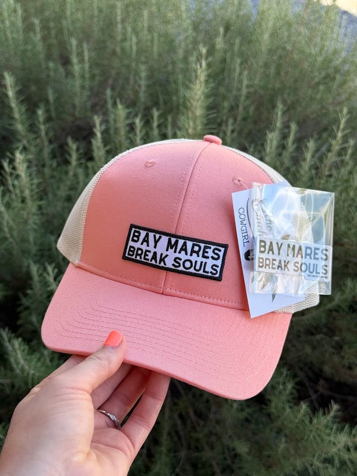 A pink trucker hat with beige mesh back panels and a black rectangular patch reading 'BAY MARES BREAK SOULS' is held by a hand outdoors in front of green plants. A branded hangtag is attached to the brim.