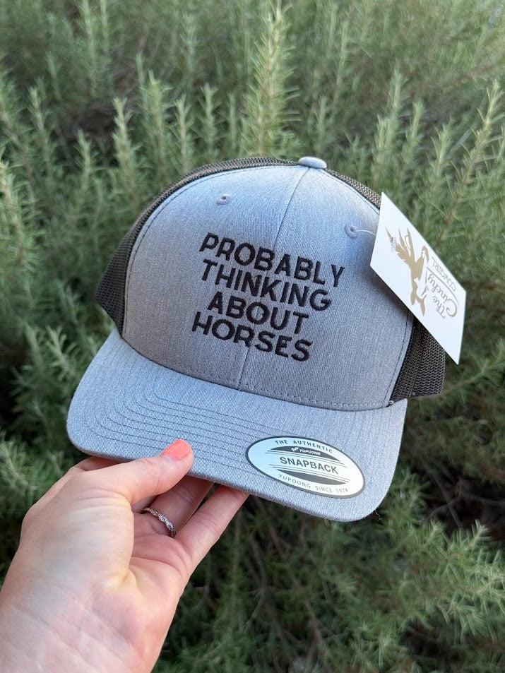 A person's hand holds a heather gray Snapback trucker hat with black mesh back panels and black embroidered text reading 'PROBABLY THINKING ABOUT HORSES'. The hat has a white hangtag and a silver Snapback sticker on the brim, set against a background of green outdoor foliage.