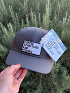 A dark gray baseball cap held by a hand, featuring a white rectangular patch with black text that reads 'COFFEE SCRIPTURE AND HORSES' and a matching paper hangtag attached. The background consists of green, textured foliage.