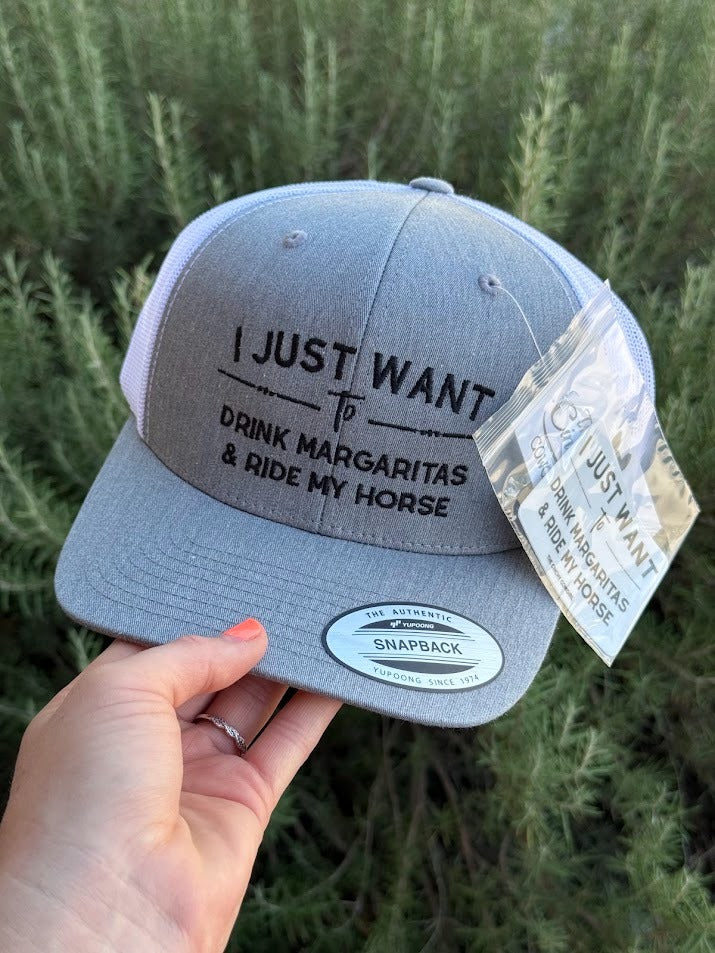 A gray snapback trucker hat with black text reading 'I JUST WANT To DRINK MARGARITAS & RIDE MY HORSE' held by a hand against a background of green plants. The hat has a gray front panel, black mesh side panels, and a white Snapback brand sticker on the brim.