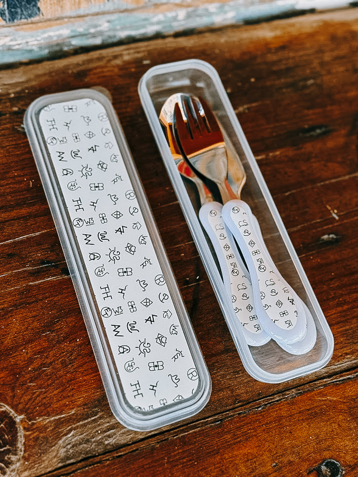 A small set of stainless steel cutlery with white handles, shown in a clear plastic case on a rustic wooden surface. The case lid and handles are printed with black symbols and text.