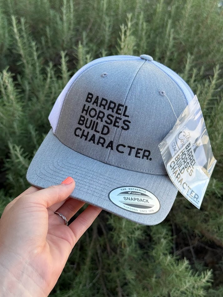 A light gray Snapback cap held by a hand, featuring black embroidered text that reads 'BARREL HORSES BUILD CHARACTER.' on the front panel, with a white mesh back and a hangtag attached.