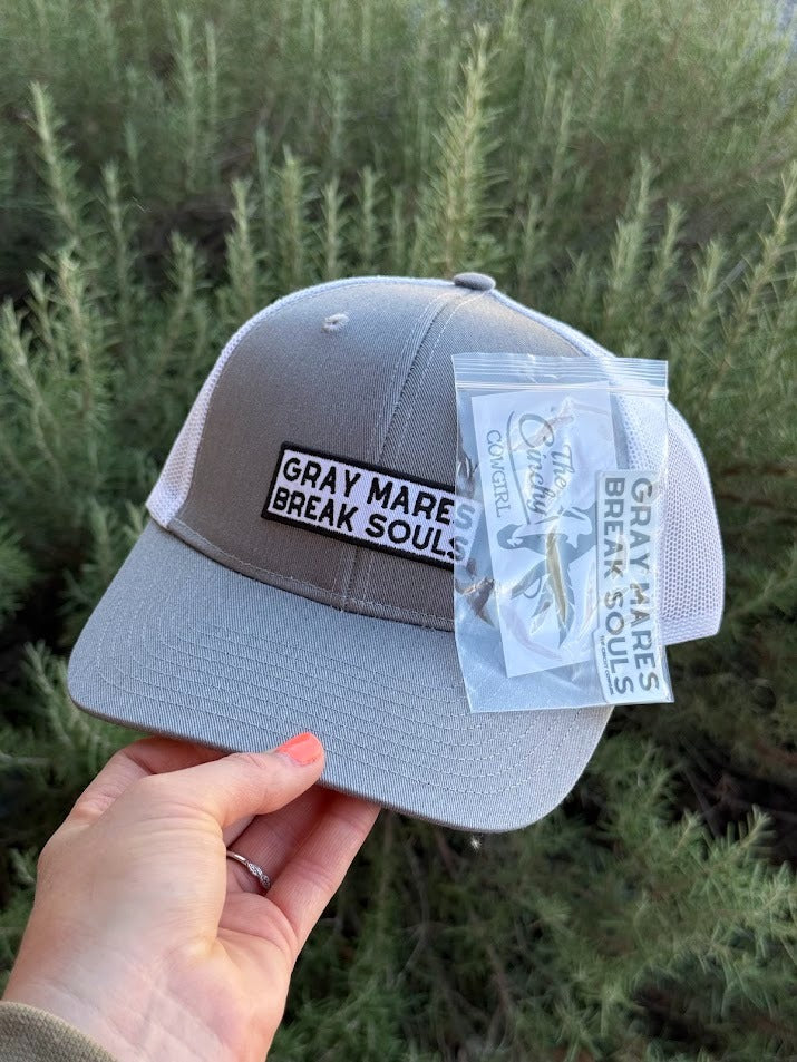 A gray trucker-style cap with white mesh side panels and a front patch reading 'GRAY MARE BREAK SOULS,' held by a hand outdoors in front of green shrubs. A clear plastic hangtag is attached to the brim.