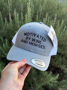 A light gray Snapback trucker hat with black embroidered text reading 'MOTIVATED BY WINE AND HORSES' on the front, held by a hand against a background of green foliage. A white hangtag is attached to the side.
