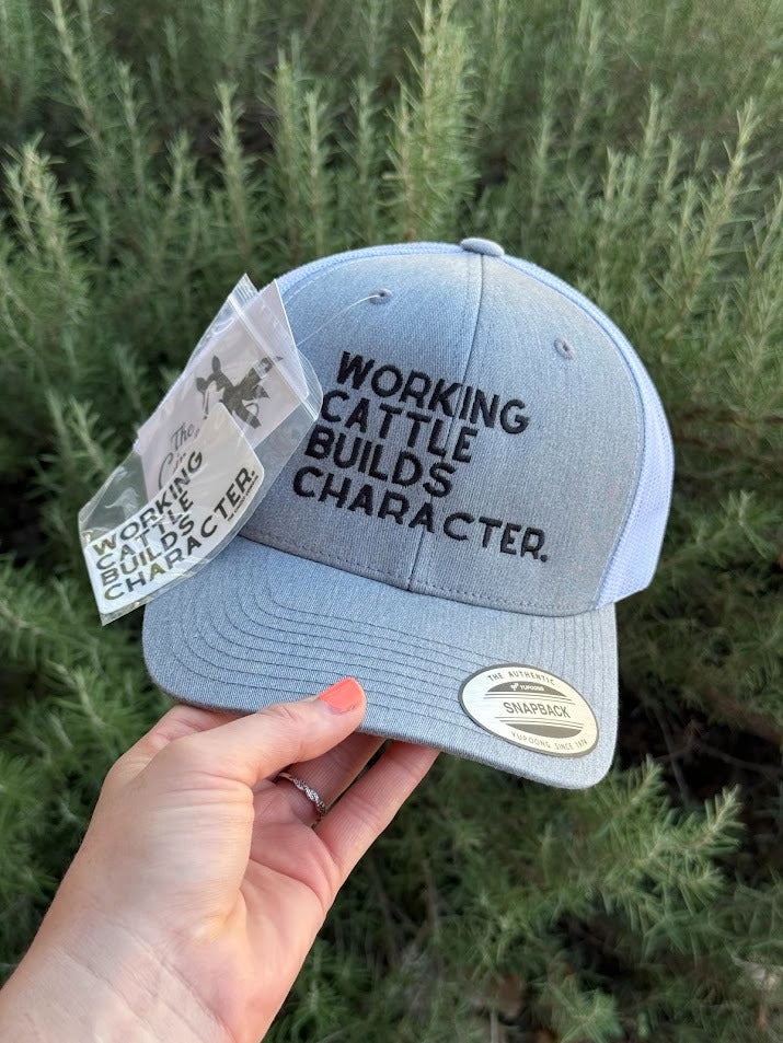 A light blue heathered Snapback cap with black embroidered text reading 'WORKING CATTLE BUILDS CHARACTER.' held by a hand against a background of green shrubs.