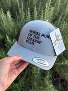 A light heather gray snapback trucker hat held by a hand, featuring black embroidered text that reads 'HORSE MOM OF THE FREAKIN' YEAR.' on the front panel.