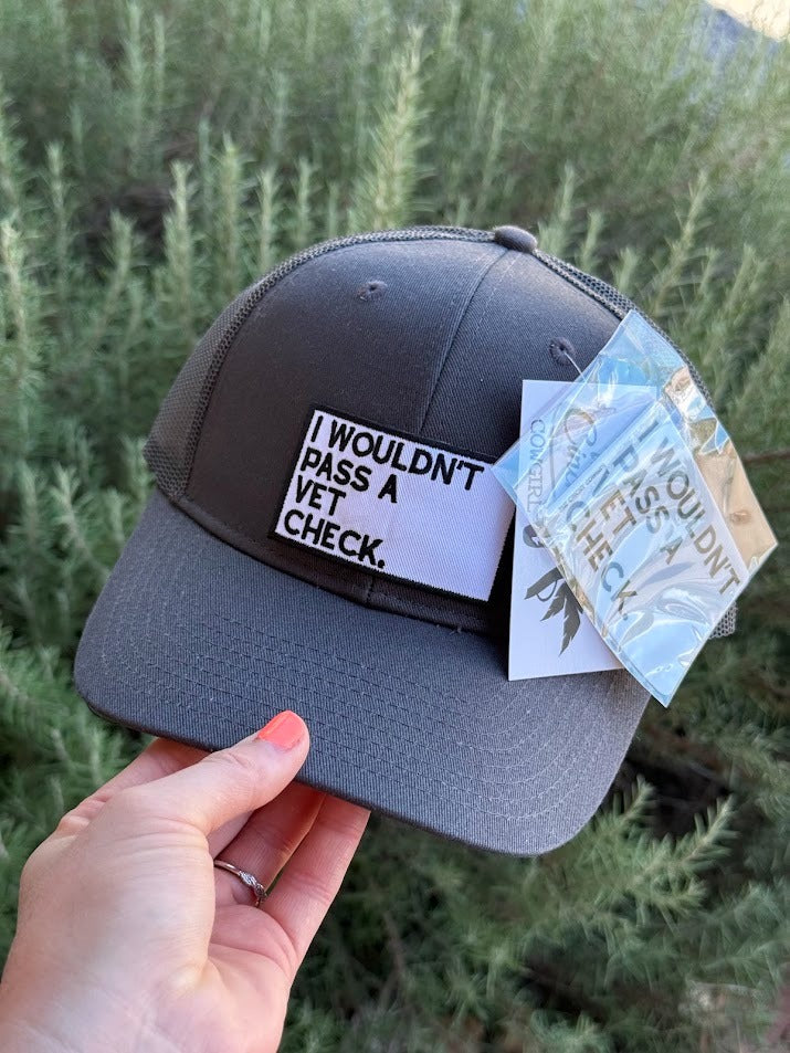 A dark gray trucker-style cap held by a hand, featuring a white patch that reads 'I WOULDN'T PASS A VET CHECK.' in black text.