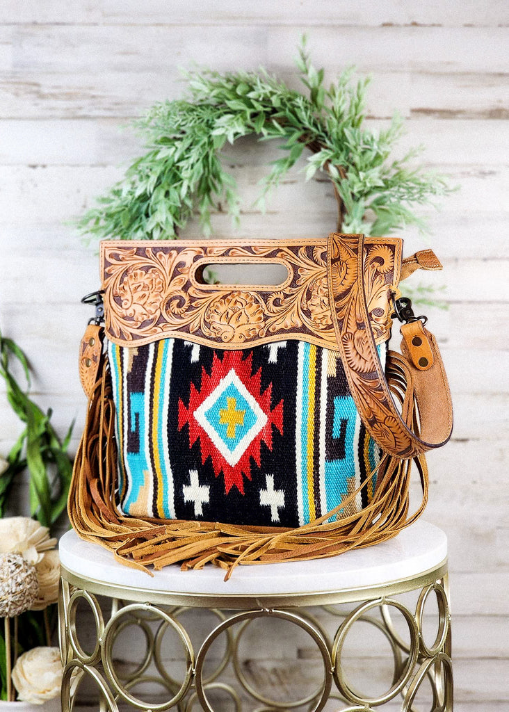 Southwestern saddle blanket purse with black, teal, and red tribal design. Tooled leather top paneling with matching strap. Fringe hands from the side of the purse.