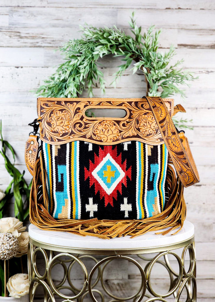Southwestern saddle blanket purse with black, teal, and red tribal design. Tooled leather top paneling with matching strap. Fringe hands from the side of the purse.
