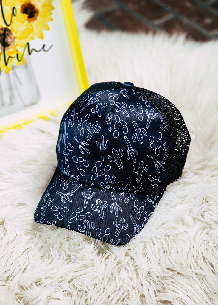 Black mesh trucker hat with white printed cacti design on front. Laying on faux rug. 