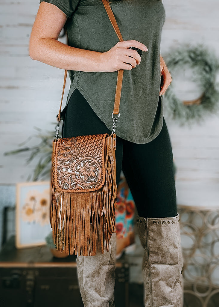 Brown Floral Tooled Crossbody Handbag on model wearing an olive green shirt, black leggings and tan knee high boots, background has western and farmhouse decor