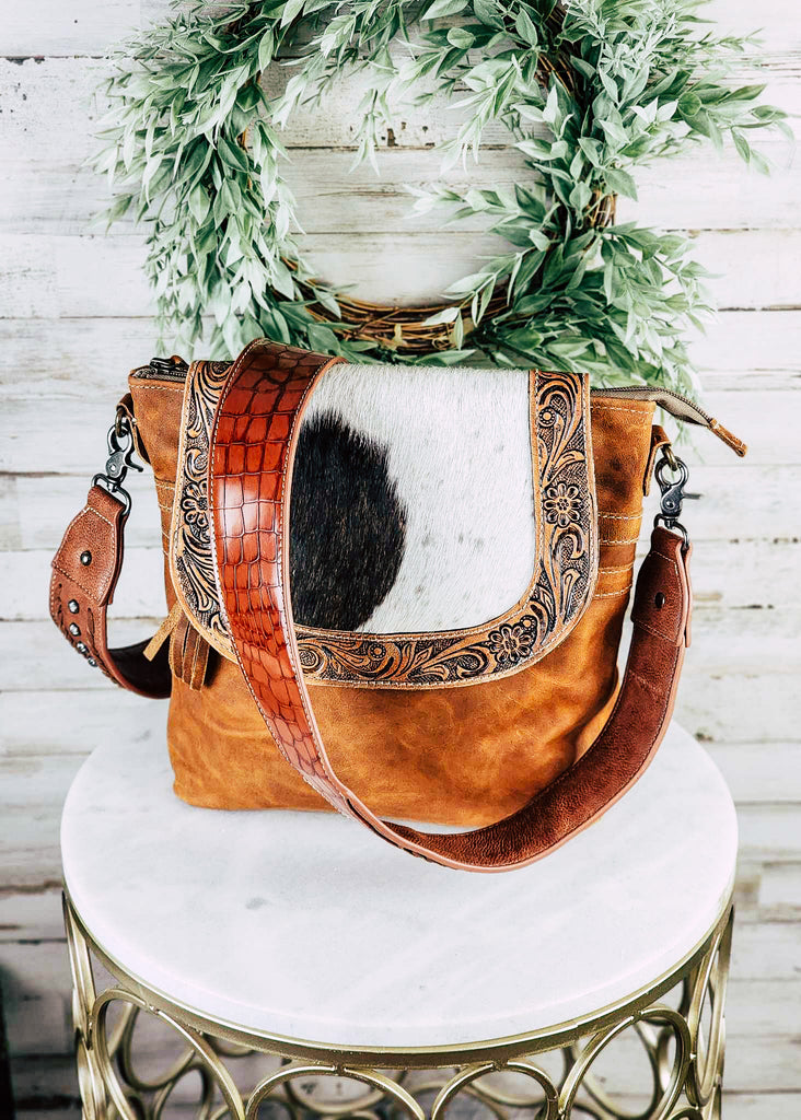 Brown gator  leather guitar or purse strap with silver concho studs and whip stitch by ends of strap. Strap is displayed connected to a leather purse with a cowhide flap with floral tooling. Both sit on top of a marble table. 