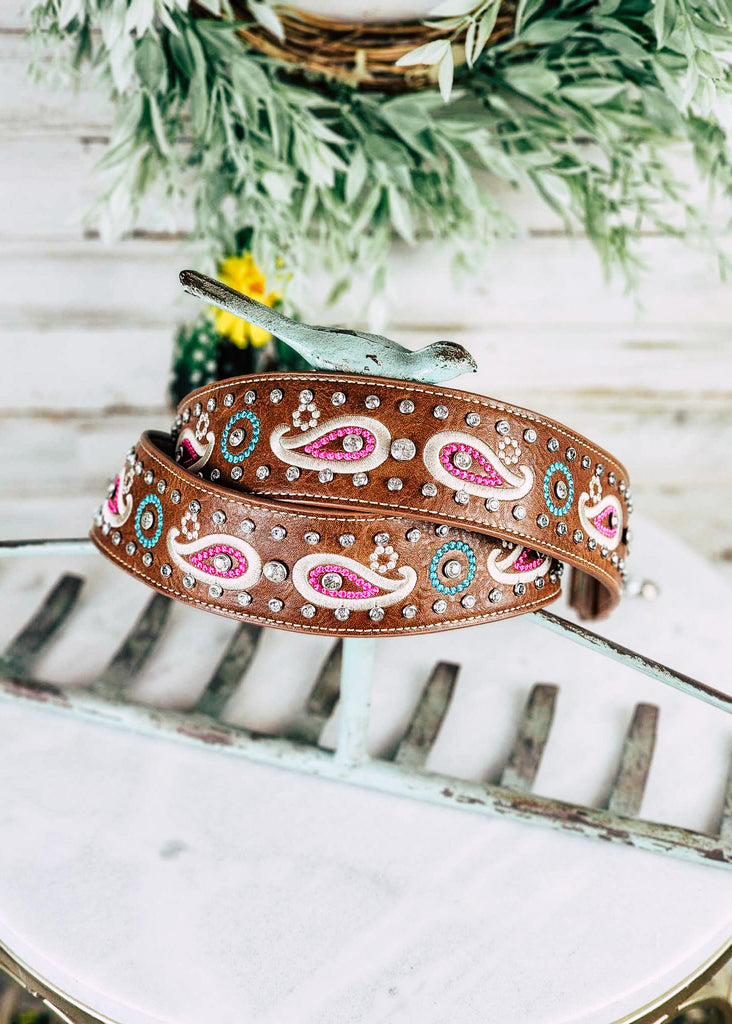 Brown leather purse or guitar strap with paisley stitching. Jewel studs line the strap and have detail inside the paisleys. Purse strap is wrapped around blue metal rake stand.
