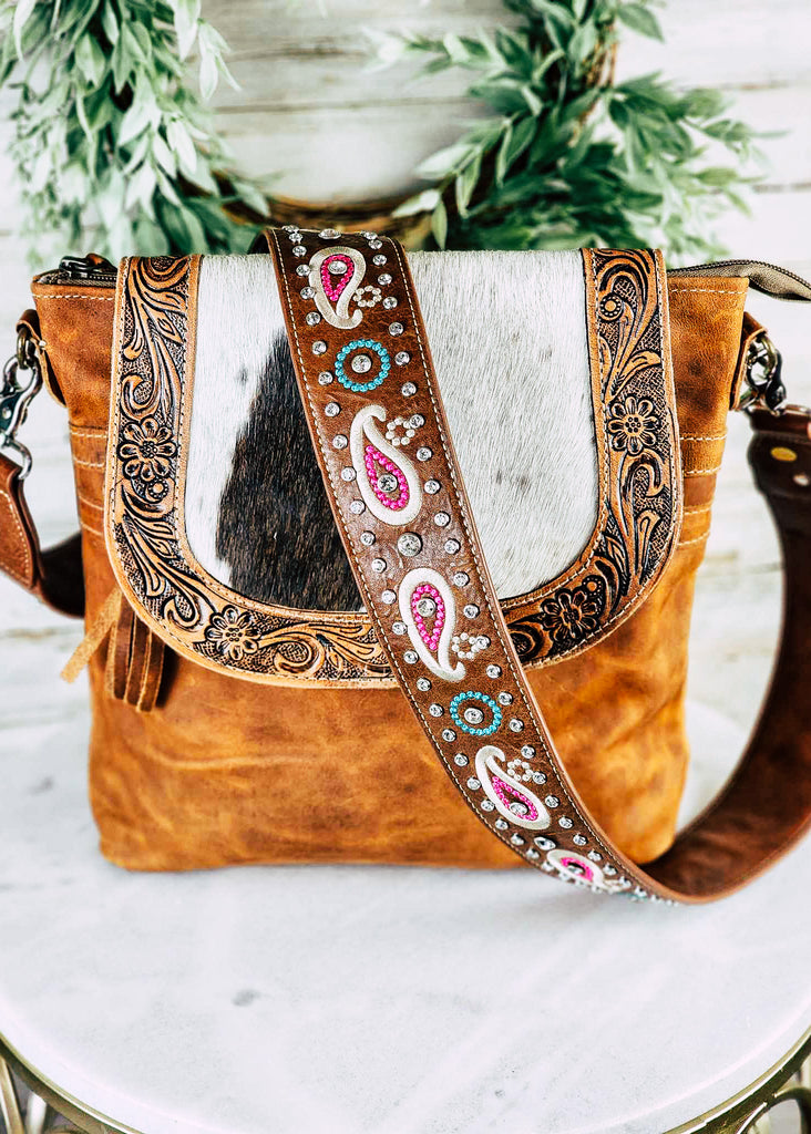 Brown leather purse or guitar strap with paisley stitching. Jewel studs line the strap and have detail inside the paisleys. Strap is connected to a brown leather purse with a cowhide and tooled leather flap. Both sits on a marble table.