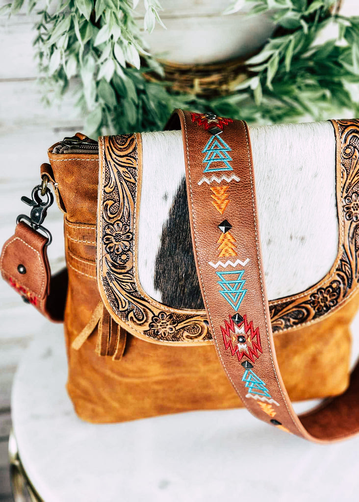 Brown southwestern embroidered guitar or purse strap with silver stud accents line the middle of the strap between embroidered design.  Strap is attached to a leather purse with floral tooling and cowhide flap. 