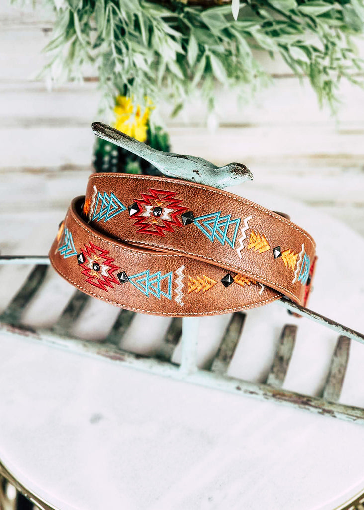 Brown southwestern embroidered guitar or purse strap with silver stud accents line the middle of the strap between embroidered design. Strap is coiled on a blue metal rake. 