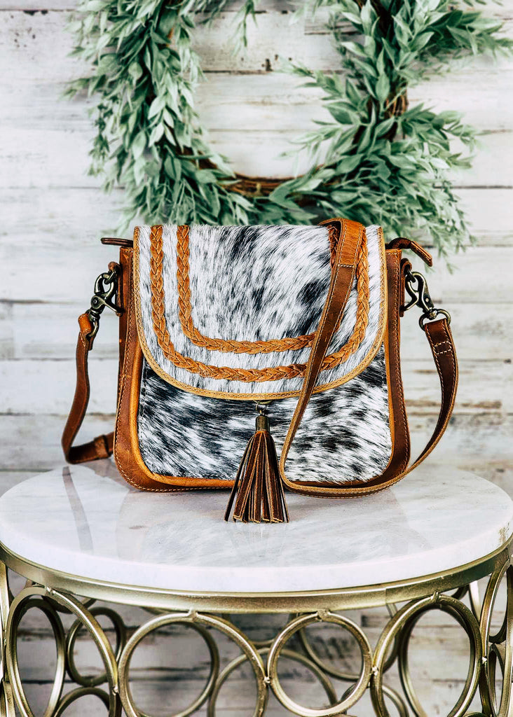 Western cowgirl purse with cowhide front and flap with braided leather accents on the flap. Adjustable shoulder strap hangs over purse with fringe accent hanging from the front of the flap. 