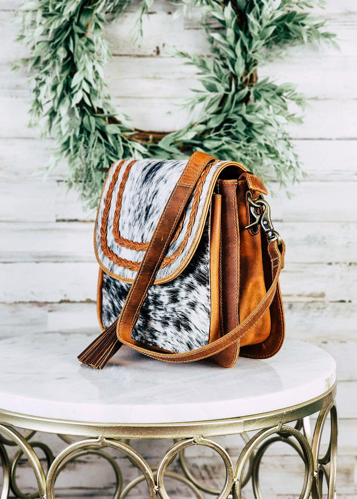 Western cowgirl purse with cowhide front and flap with braided leather accents on the flap. Adjustable shoulder strap hangs over purse with fringe accent hanging from the front of the flap.