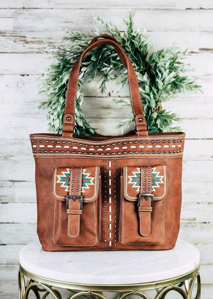 Concealed carry with aztec embroidered dual pocket with buckle. Leather purse with whip stitch and silver studs decor. Matching leather handles. Purse sits on marble table top.