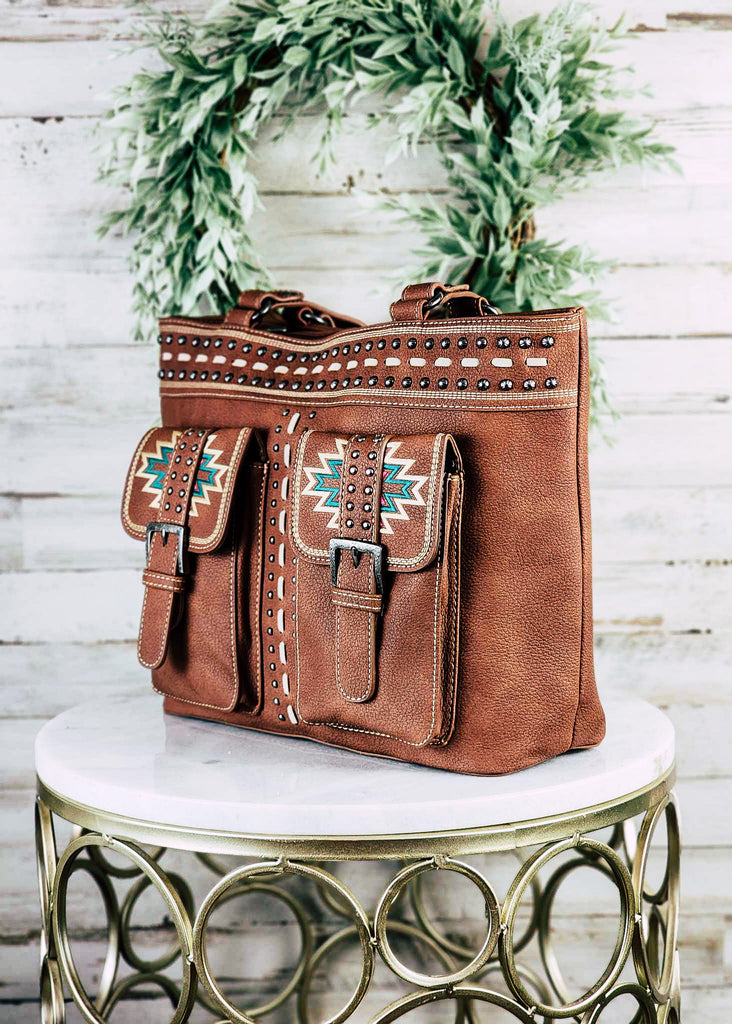 Concealed carry with aztec embroidered dual pocket with buckle. Leather purse with whip stitch and silver studs decor. Matching leather handles. Purse sits on marble table top.