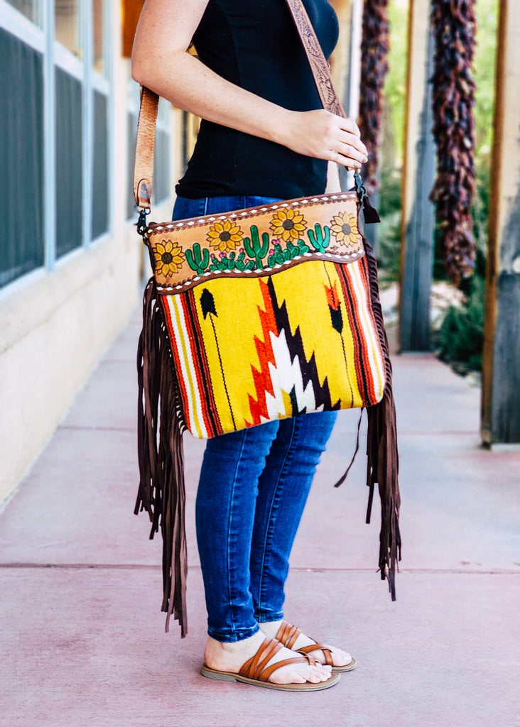 Concealed carry saddle blanket bag with fringe on the sides of the bag. Tooled cactus and sunflower leather paneling on the top of the purse with white buck stitch. Model wears this with tooled shoulder strap. 
